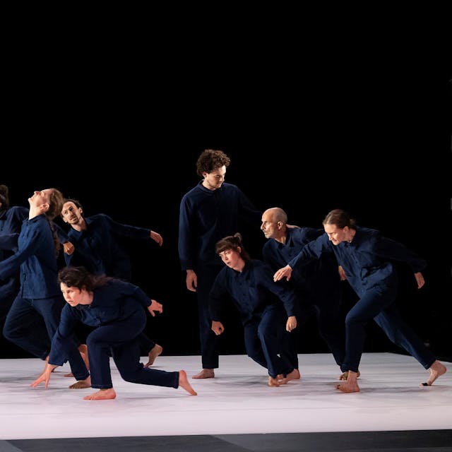 Die Tänzerinnen und Tänzer tragen blaue Hosen und Hemden und bewegen sich alle auf eine Tänzerin zu, die ihnen entgegenschaut. Der Hintergrund ist schwarz, der Bühnenboden ist weiß.&nbsp;