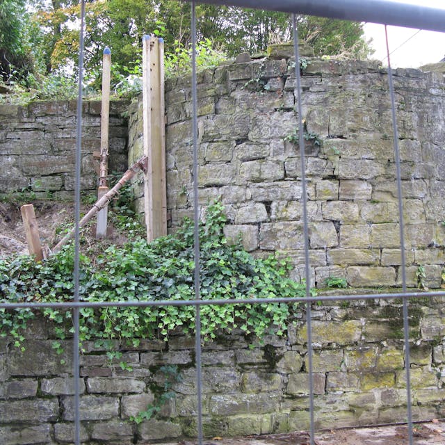 Im Vordergrund steht ein Bauzaun. Dahinter ist die halbrunde Apsis der zerstörten Synagoge zu sehen. Die Bruchsteinmauern sind durch Stützen gesichert.