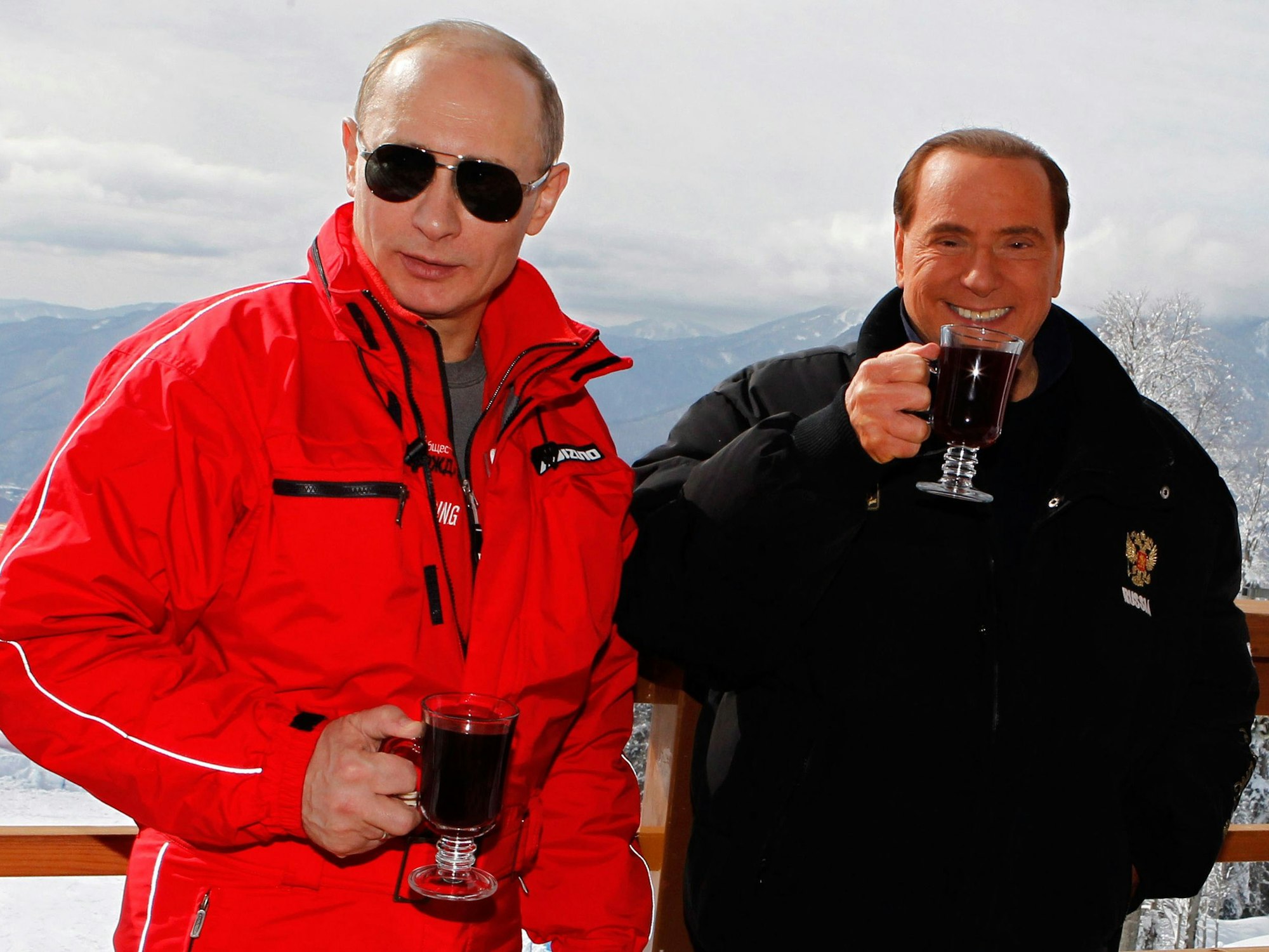 Wladimir Putin im roten Ski-Anzug steht neben Silvio Berlusconi. Beide halten ein Getränk in der rechten Hand.