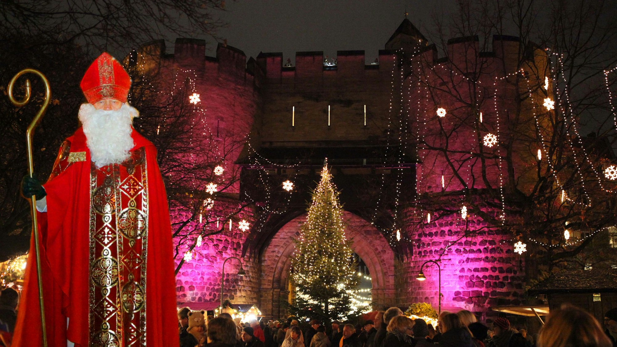 Ein Nikolaus in vollem Ornat steht vor der Kulisse der festlich geschmückten Torburg, ein Weihnachtsmarkt steht unter dem Torbogen.