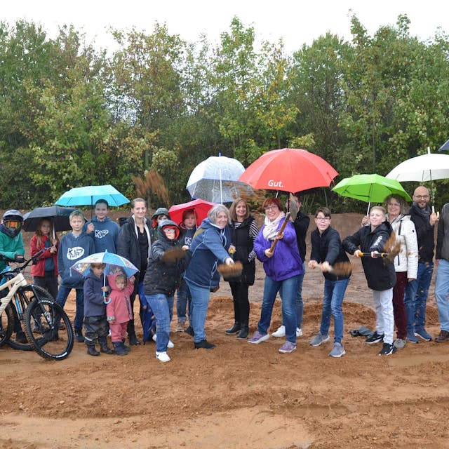 Eine größere Gruppe Menschen, Kinder und Erwachsene, steht mit Regenschirmen auf einem Bauplatz. Einige haben mit einem Spaten einen symbolischen Spatenstich vollzogen.