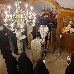 Orthodoxe Priester und Nonnen stehen in einer Kapelle unter einem Kronleuchter. An den holzvertäfelten Wänden sind sakrale Gemälde zu sehen.
