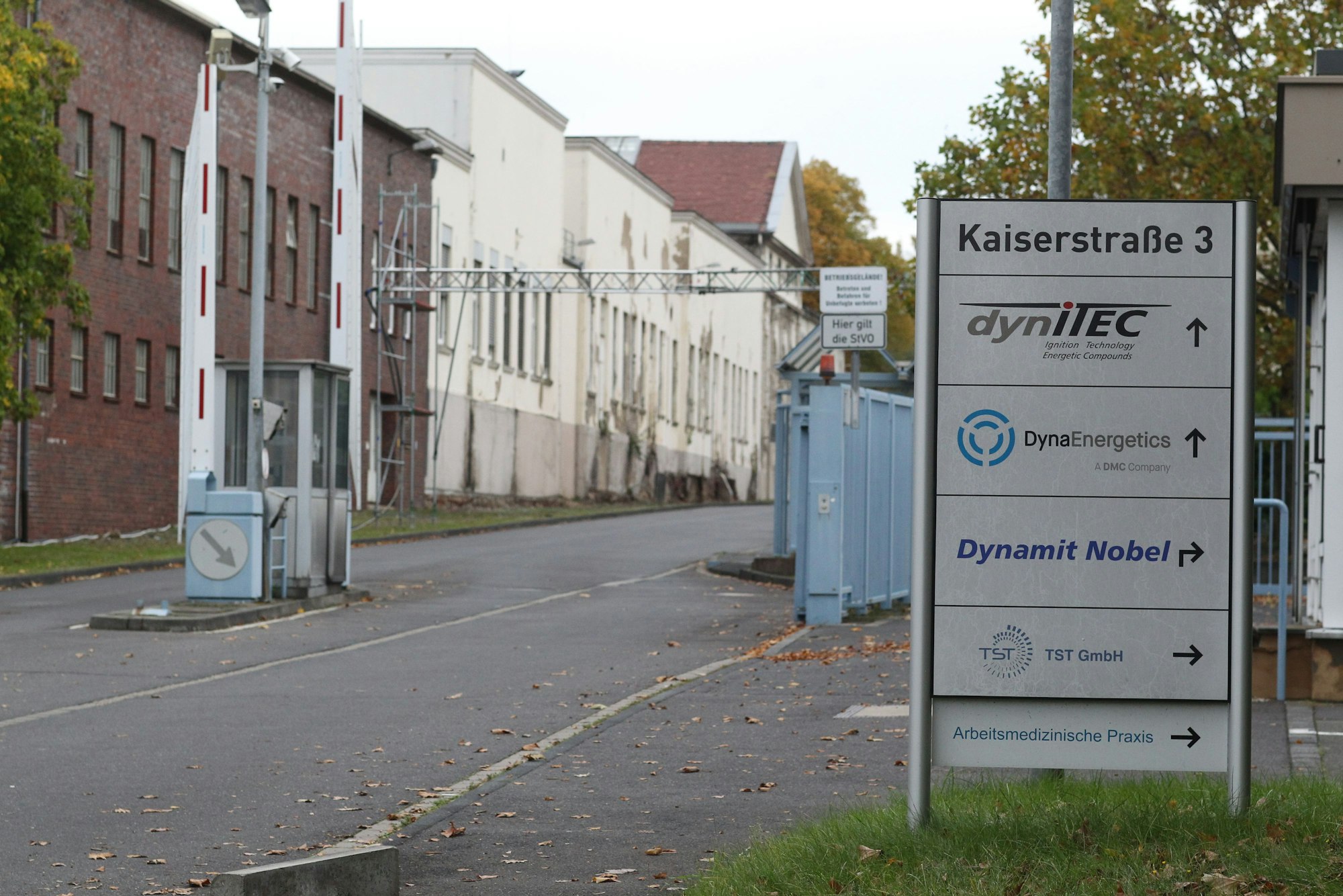 Die Zufahrt zu einem Industriegelände. Links stehen Fabrikgebäude, rechts eine Informationssäule mit den Namen ansässiger Firmen.