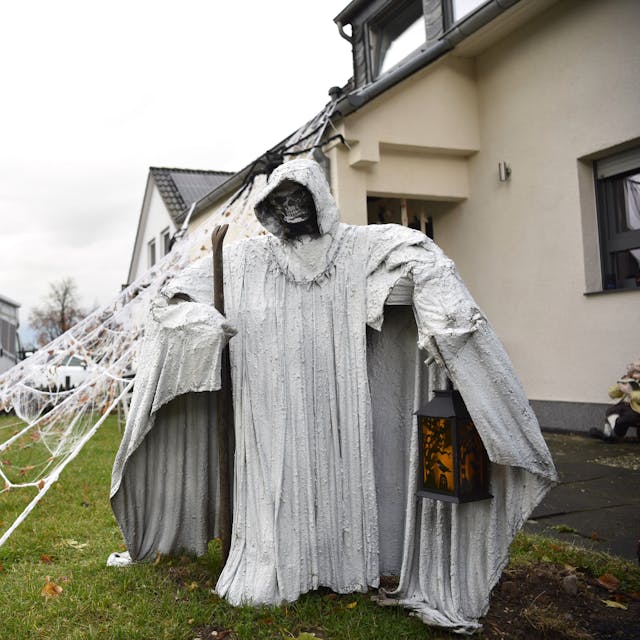 Eine Figur in einem langen weißen Mantel, mit Kapuze, Sense und Laterne steht vor einem Haus. Im Hintergrund sind künstliche Spinnweben, Skelette und Kürbisse zu erkennen.