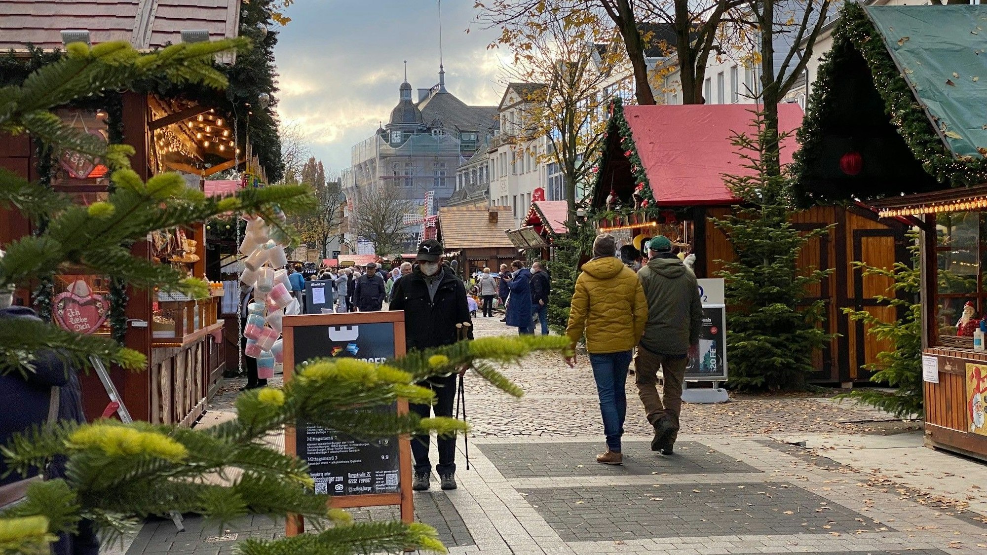 Weihnachtsmarktbuden aus Holz mit geschmückten Dächern reichen sich über den Marktplatz. Menschen schlendern zwischen ihnen umher.
