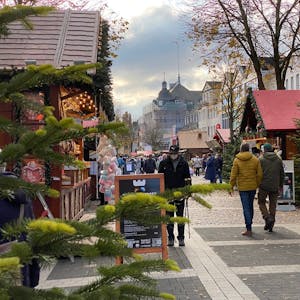 Weihnachtsmarktbuden aus Holz mit geschmückten Dächern reichen sich über den Marktplatz. Menschen schlendern zwischen ihnen umher.