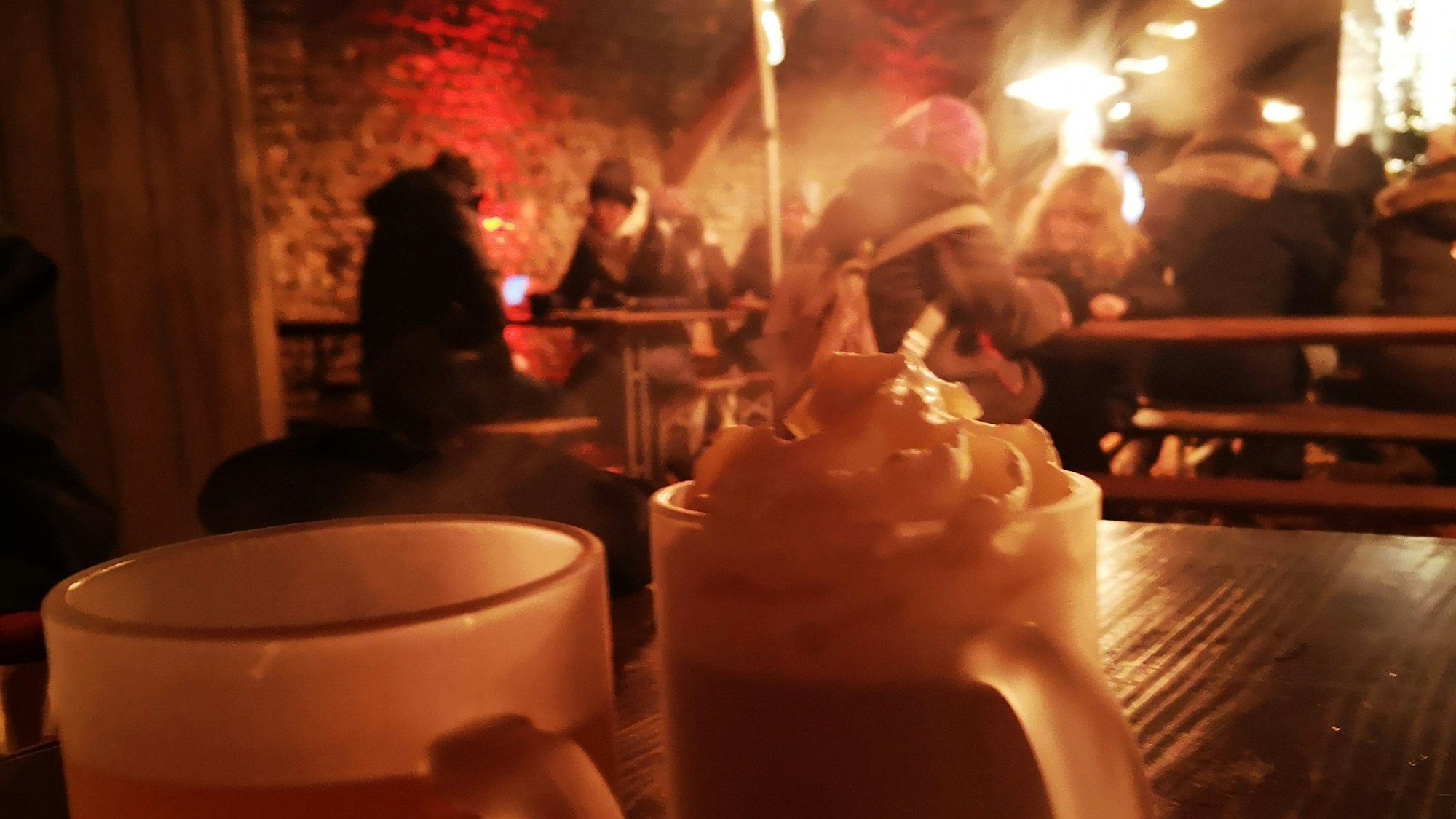 Auf einem Holztisch stehen zwei dampfende Tassen mit Punsch und Sahne-Kakao. Im Hintergrund sitzen Menschen mit Winterjacken an Biertischen und -bänken.