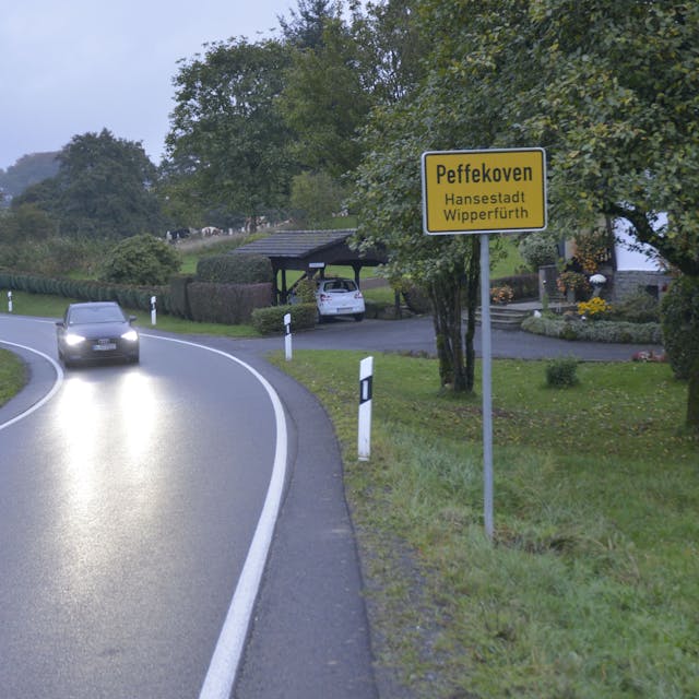 Das Ortsschild von Wipperfürth-Peffekoven steht am Rande einer Straße, ein Auto fährt mit eingeschalteten Scheinwerfern daran vorbei.