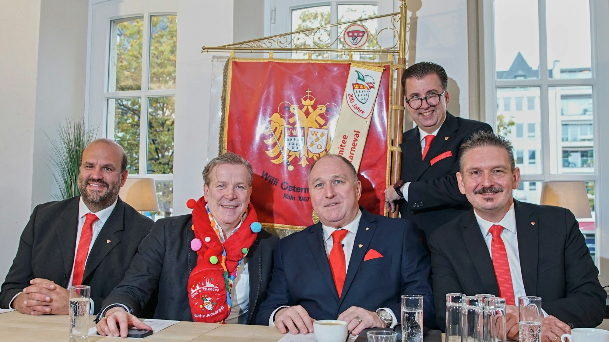 Fünf Männer in Anzügen sitzen an einem Tisch und lächeln in die Kamera.