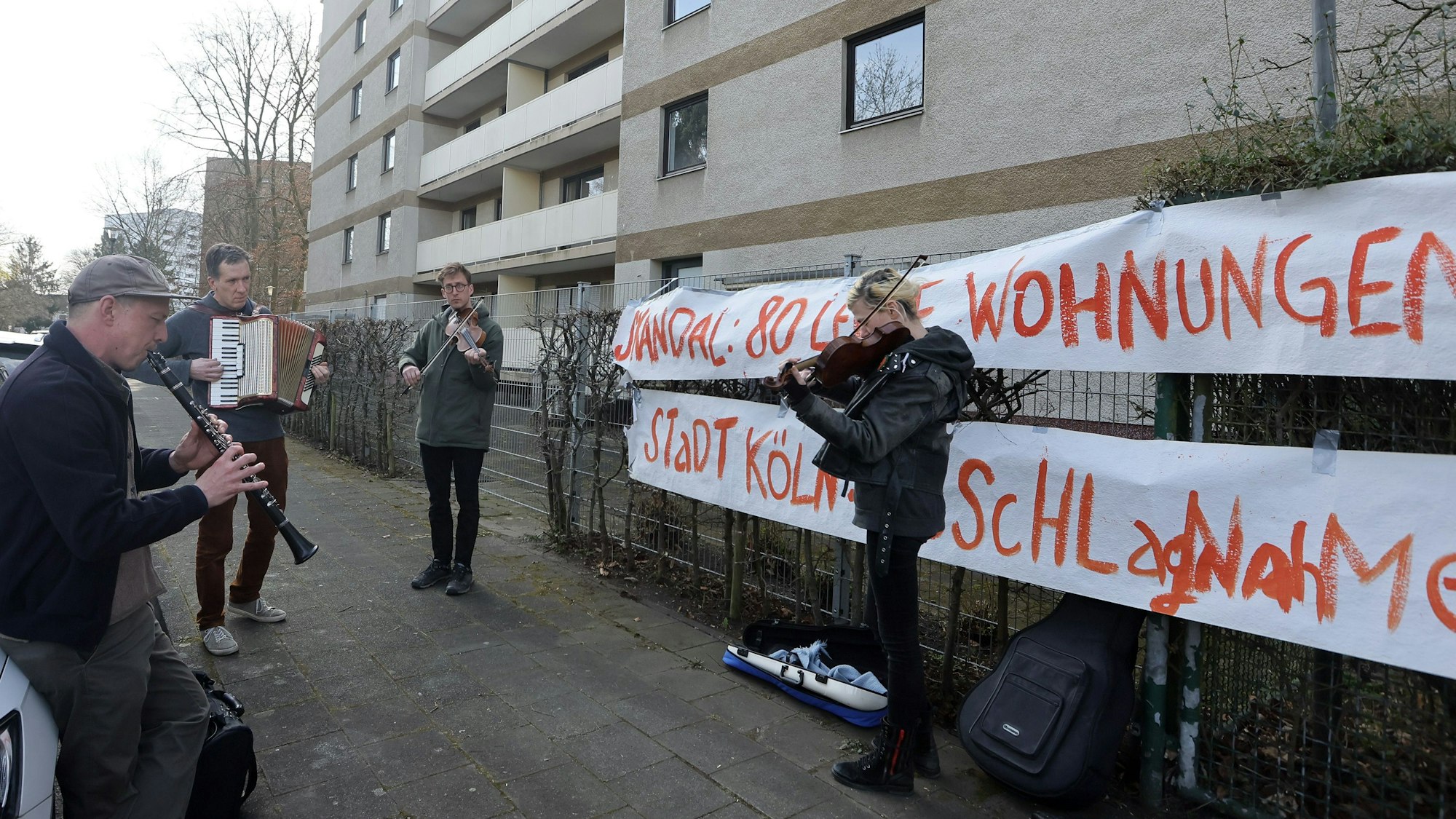 Musiker stehen vor einem Häuserblock, am Zaun vor dem Gebäude hängt ein Protest-Banner mit der Aufforderung an die Stadt, 80 leere Wohnungen zu beschlagnahmen.
