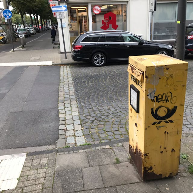 Ein total rostiger Briefkasten steht auf dem Bürgersteig an einer Straße.
