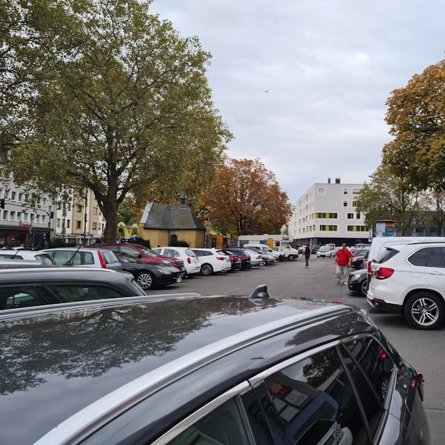 Ein langgezogener Platz mit vielen parkenden Autos und einigen Bäumen am Rand ist zu sehen. Auch eine kleine Kapelle steht am linken Rand der Platzfläche.