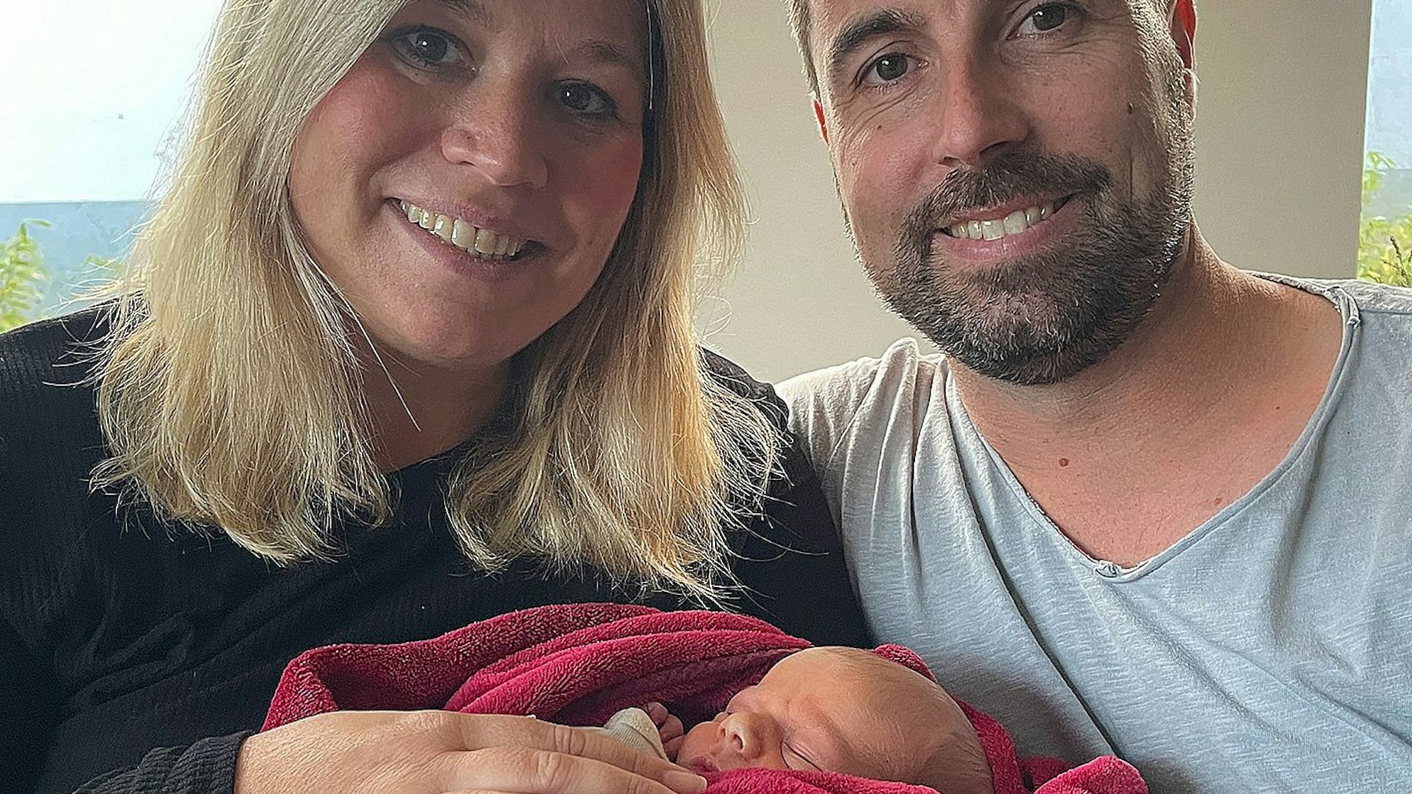 Jennifer und Krishan Meuren halten ihren neugeborenen Sohn Levi in einer roten Decke im Arm.