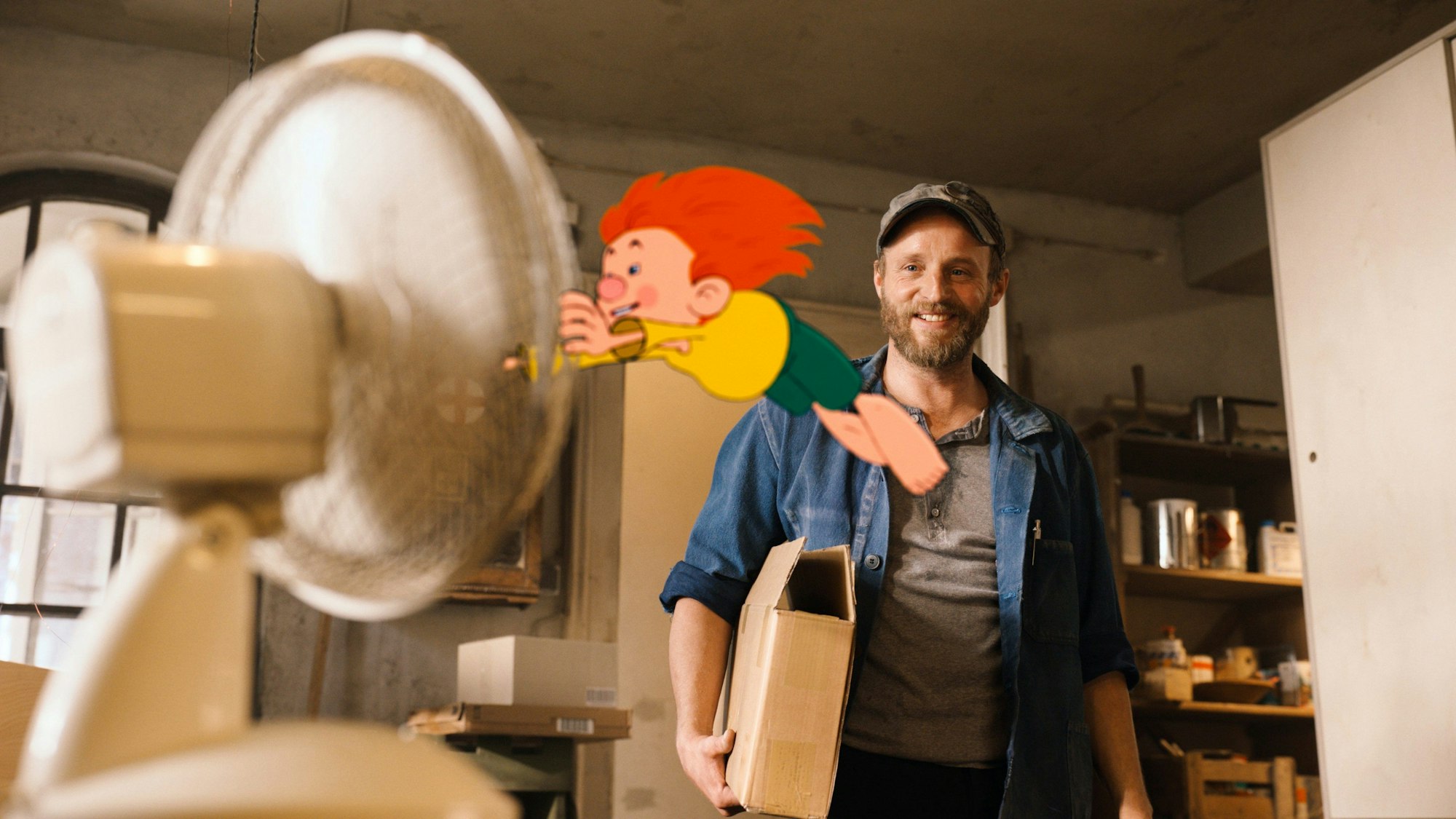 Florian Brückner als Florian, Neffe von Meister Eder, steht mit einer Kappe in einer Szene des Films „Neue Geschichten vom Pumuckl“ in der Werkstatt, während der Pumuckl sich an einem Ventilator festhält, der ihn wegbläst.