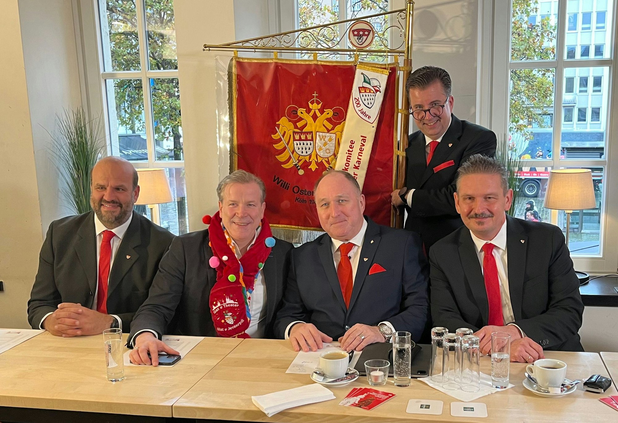 Vier Männer sitzen an einem Tisch, hinter ihnen steht ein weiterer und hält die Fahne der Willi-Ostermann-Gesellschaft.