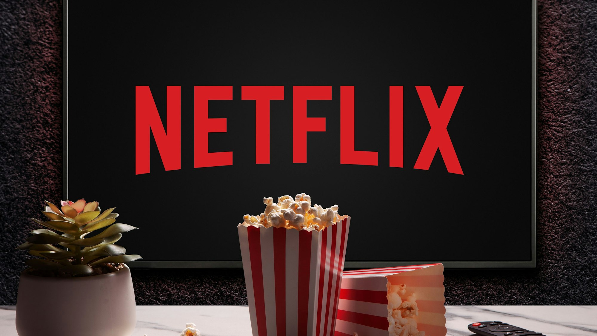 Hier zu sehen: Ein Bildschirm mit dem roten Logo von Netflix hängt an einer Wand. Im Vordergrund sind zwei Tüten Popcorn, eine Fernbedienung und eine Pflanze zu sehen.