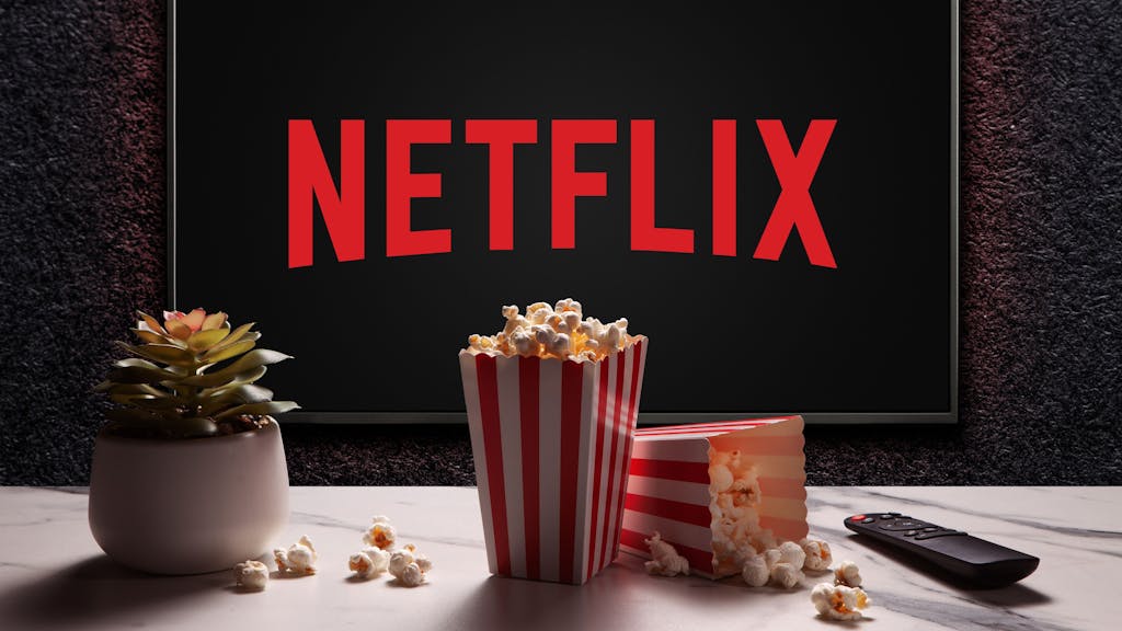 Hier zu sehen: Ein Bildschirm mit dem roten Logo von Netflix hängt an einer Wand. Im Vordergrund sind zwei Tüten Popcorn, eine Fernbedienung und eine Pflanze zu sehen.