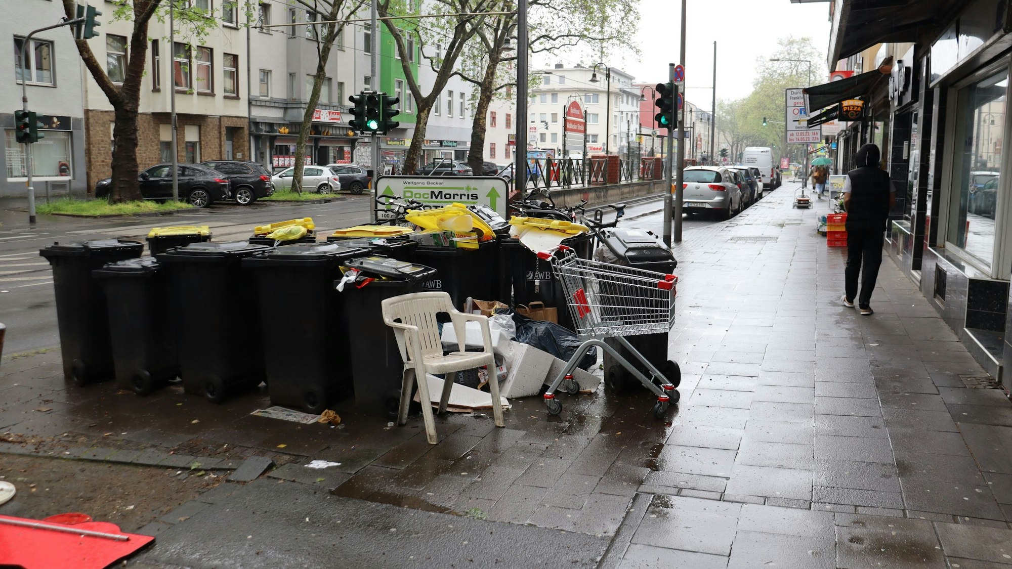 Auf einem Bürgersteig an einer belebten Straße mit Stadtbahn-Haltestelle stehen viele Mülltonnen, daneben weggeworfene Plastikstühle und ein leerer Einkaufswagen.