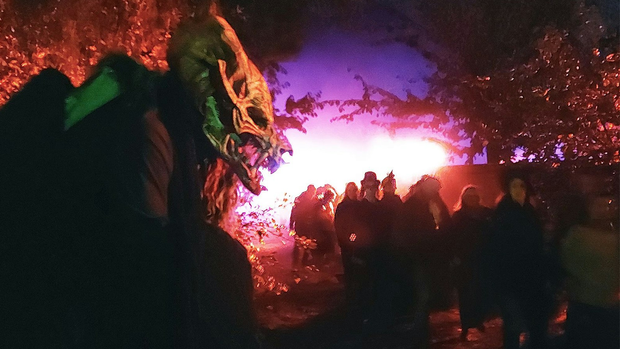 Vorne lauert das maskierte Ungeheuer, während eine Besucherschar vorbeispaziert. Die Szenerie ist in rotes und blaues Licht getaucht.