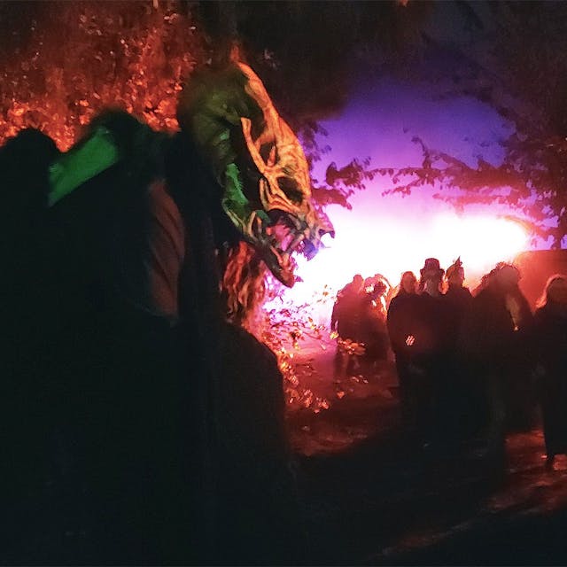 Vorne lauert das maskierte Ungeheuer, während eine Besucherschar vorbeispaziert. Die Szenerie ist in rotes und blaues Licht getaucht.