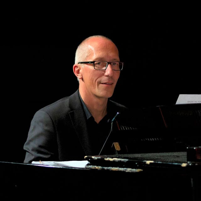 Ein Mann mit Glatze, Brille und dunklem Hemd sitzt an einem Klavier.