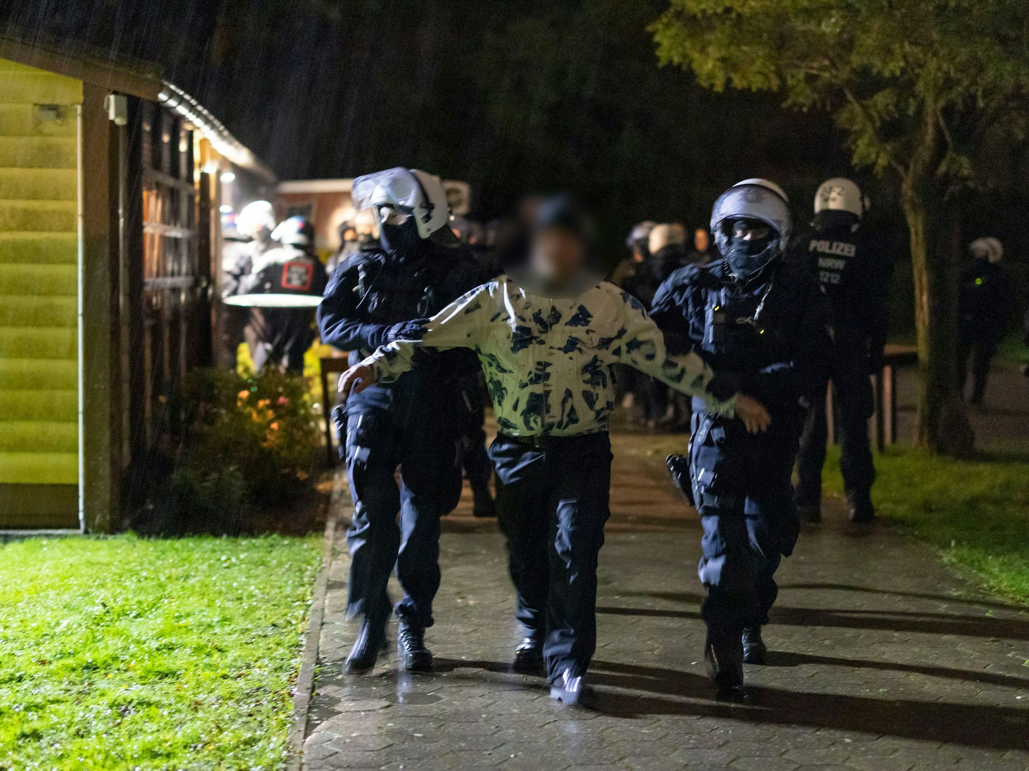 Polizeibeamte stehen am Samstag, 28. Oktober 2023, vor dem Vereinsheim einer Kleingartenanlage in Gelsenkirchen, während Teilnehmer eines mutmaßlich rechten Konzerts einzeln hinausgeführt und zur Personalienfeststellung gebracht werden.