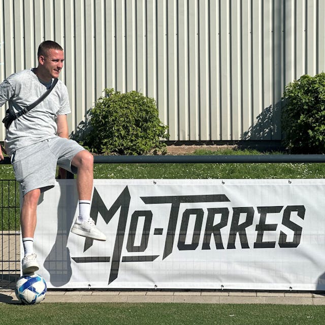 Ein junger Mann sitzt auf einem Zaun, an dem das Banner mit der Aufschrift Mo-Torres befestigt ist.