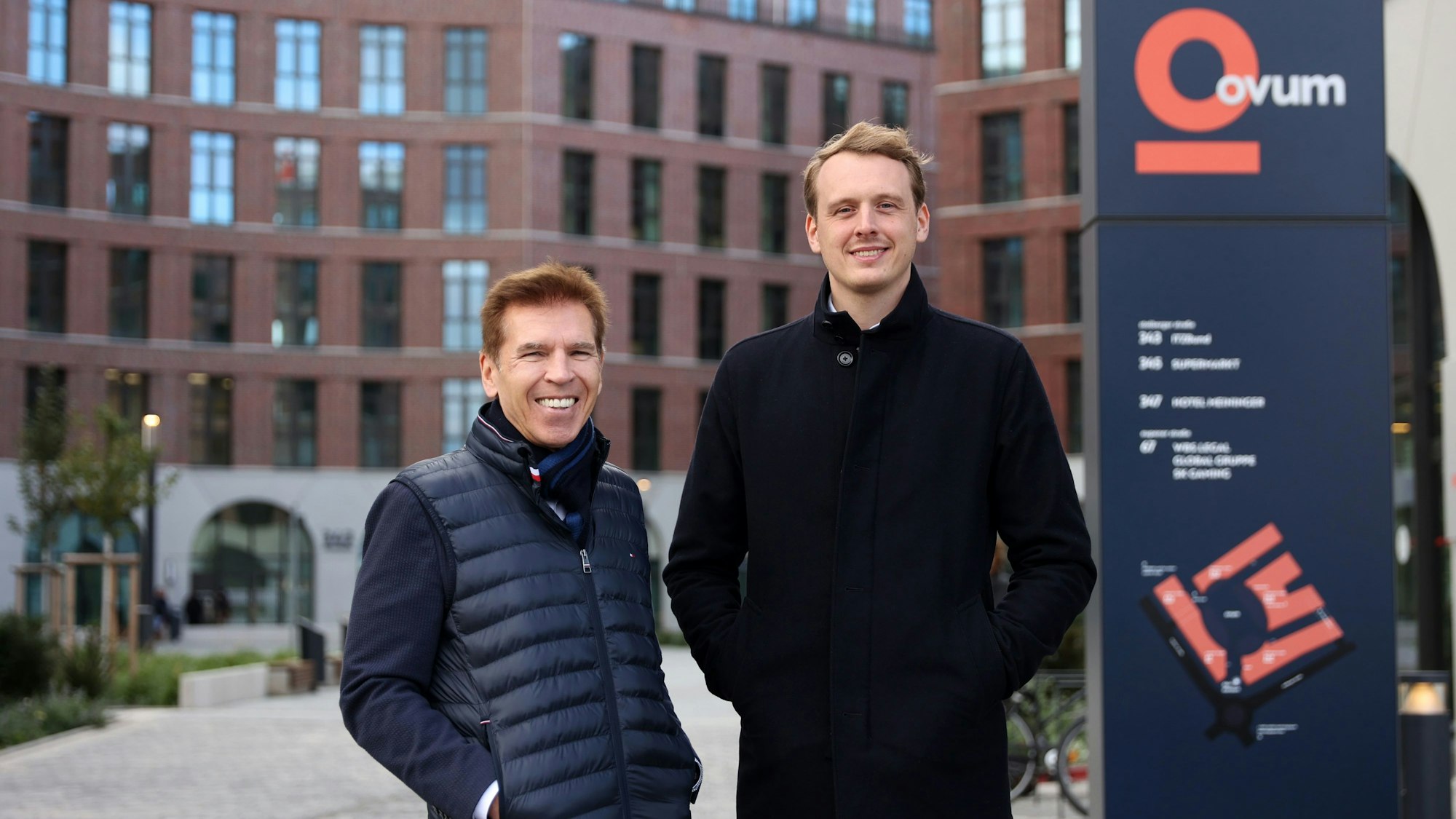 Zwei Männer stehen vor einem Gebäude und lächeln in die Kamera.