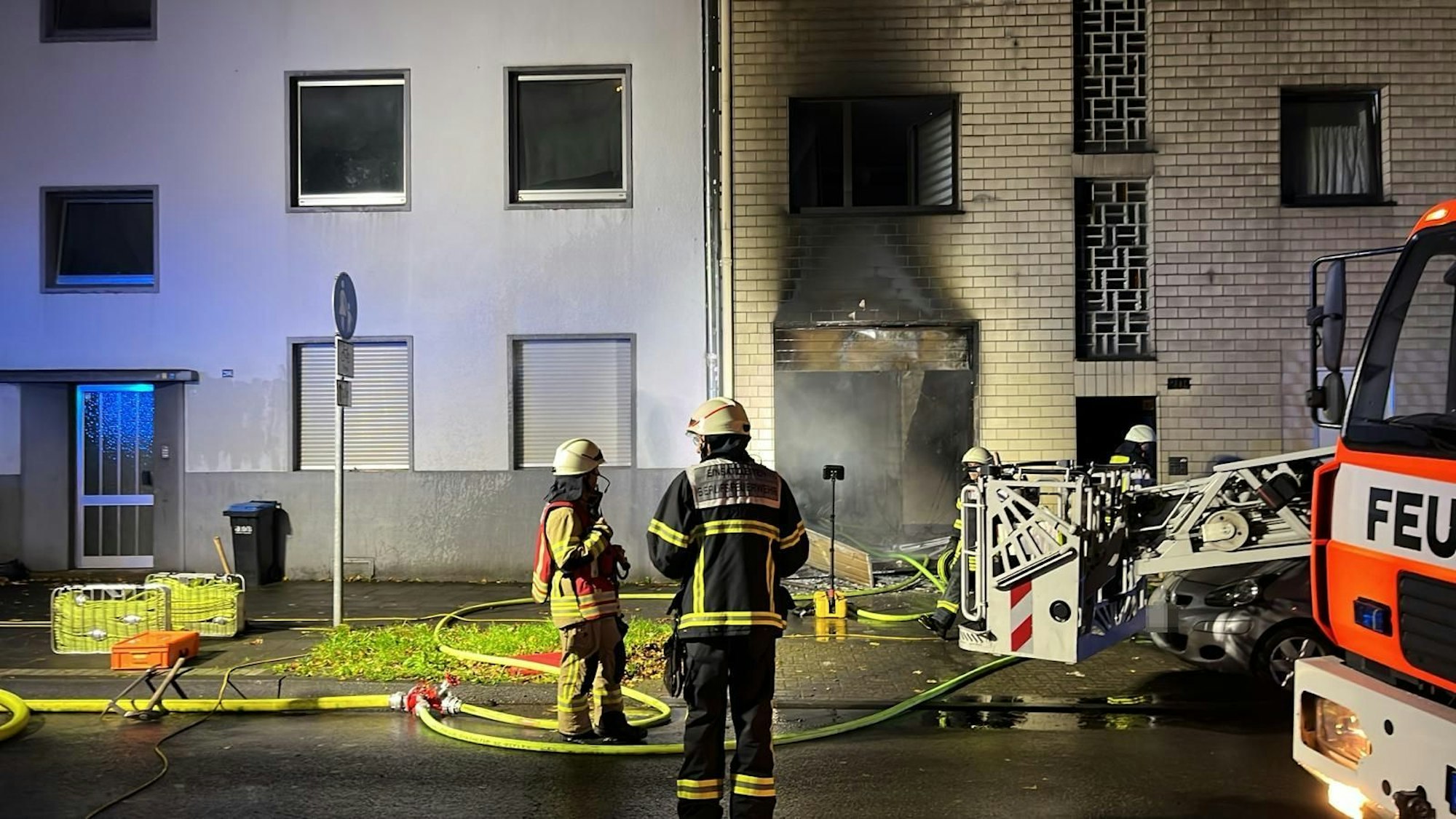 Zwei Feuerwehrmänner stehen vor einer ausgebrannten Haustür. Recht steht ein Feuerwehrwagen.