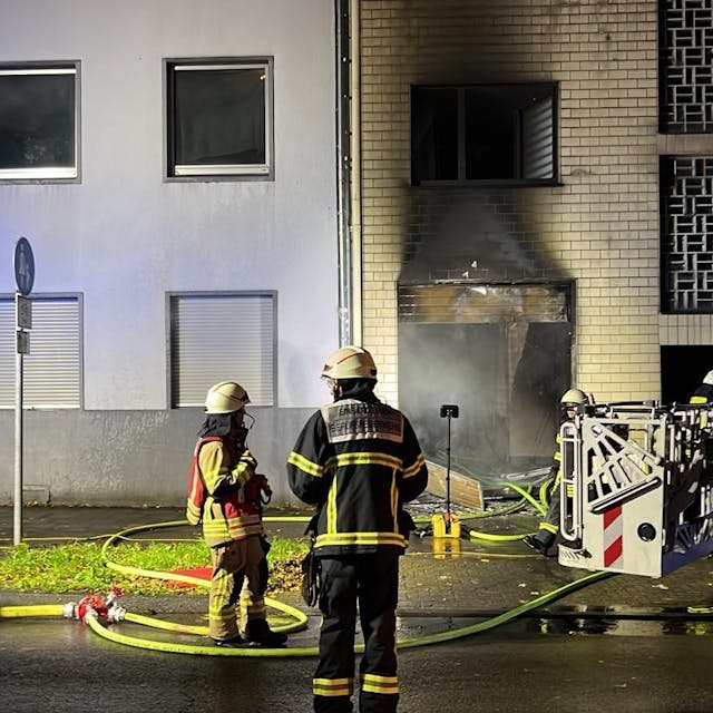 Zwei Feuerwehrmänner stehen vor einer ausgebrannten Haustür. Recht steht ein Feuerwehrwagen.