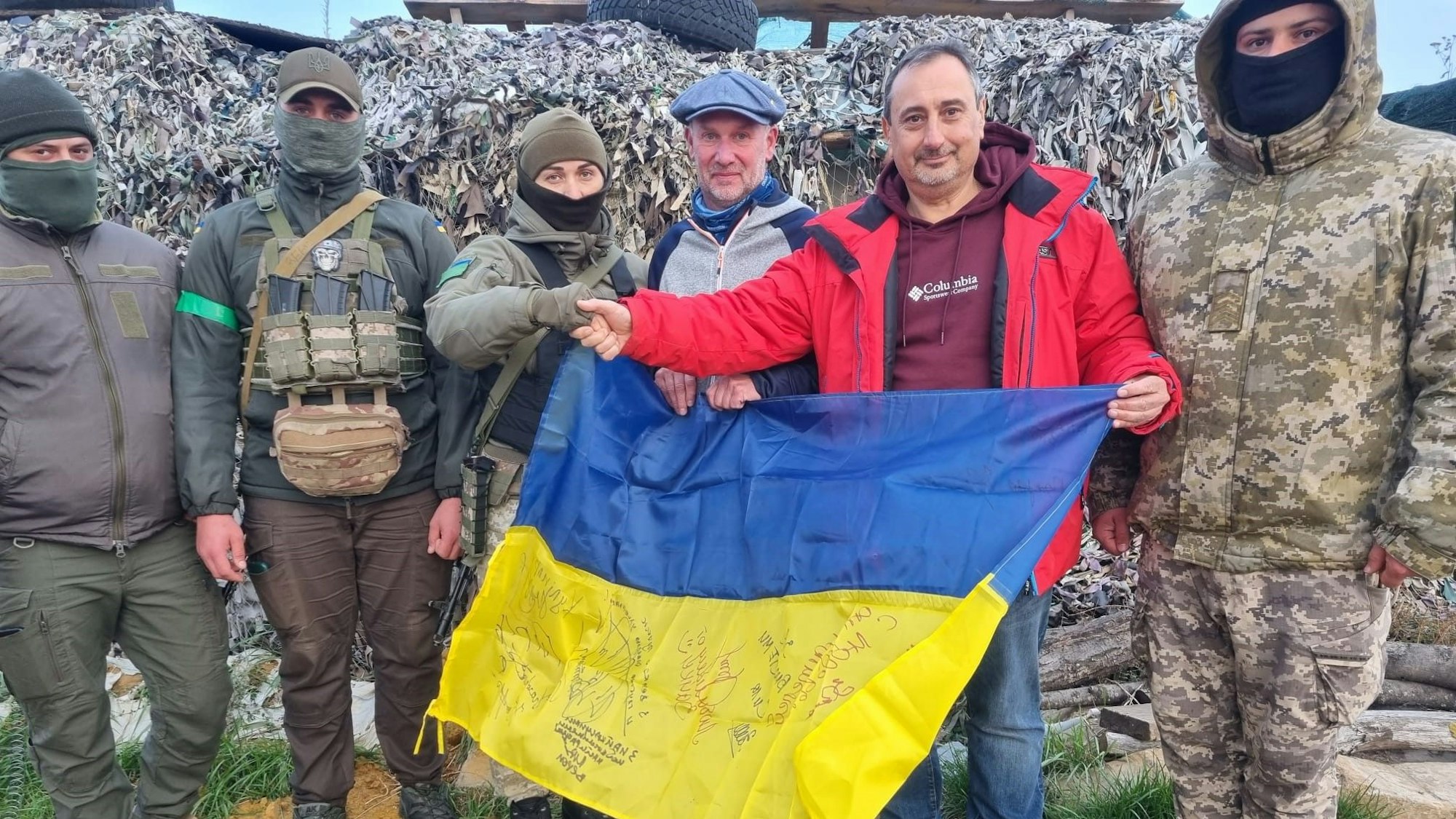 Zwei Männer stehen mit einer Ukraine-Flagge in der Hand bei an einem Kontrollposten in der Ukraine. Neben ihnen stehen mehrere vermummte Soldaten.