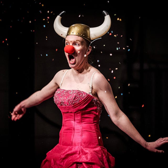 Eine Schauspielerin im roten, schulterfreien Kleid trägt eine rote Clownsnase und einen gehörnten Wikingerhelm, Szene aus „Frühlingserwachen“ im Theater der Keller