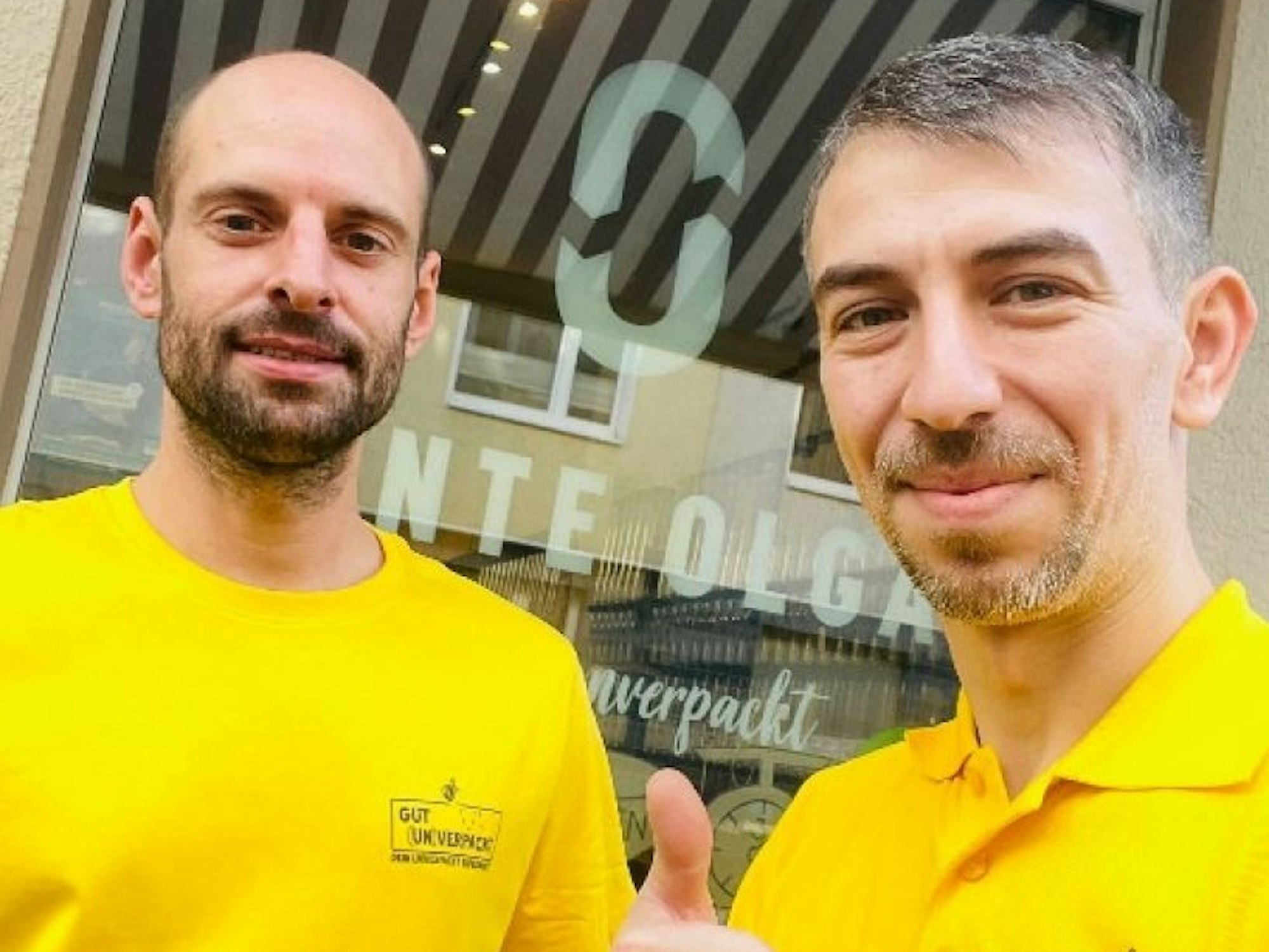 Zwei Männer in gelben Poloshirts stehen vor einem Schaufenster und schauen in die Kamera.