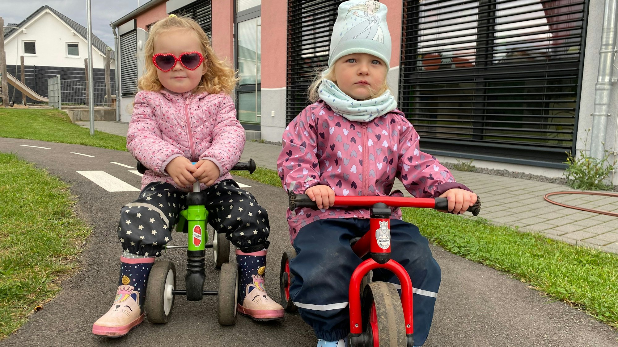 Zwei Mädchen sitzen auf Dreirädern. Eines trägt eine Sonnenbrille.