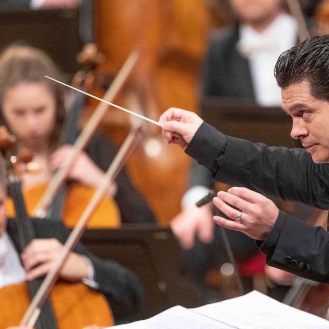 Cristian Măcelaru schaut konzentriert, hinter ihm musizieren Mitglieder des Orchesters.