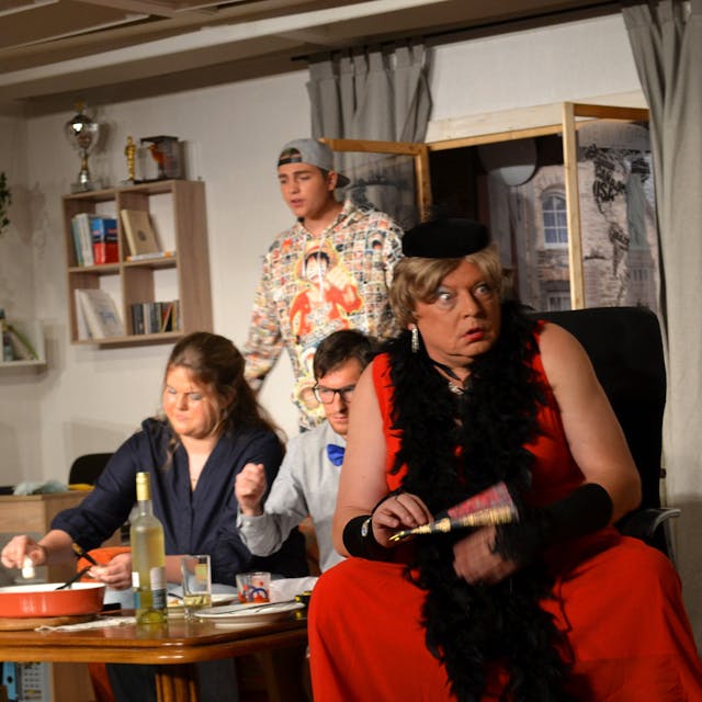 Ein Mann in einem roten Kleid mit schwarzer Federboa und Hütchen sitzt auf einem Stuhl und reißt grotesk die Augen auf, hinter ihm sind drei weitere Schauspieler.