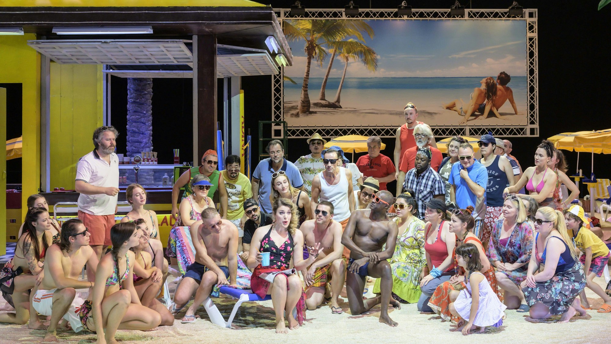 Auf der Bühne ist eine Strandszene zu sehen. Die zahlreichen Darstellerinnen und Darsteller tragen Badekleidung.