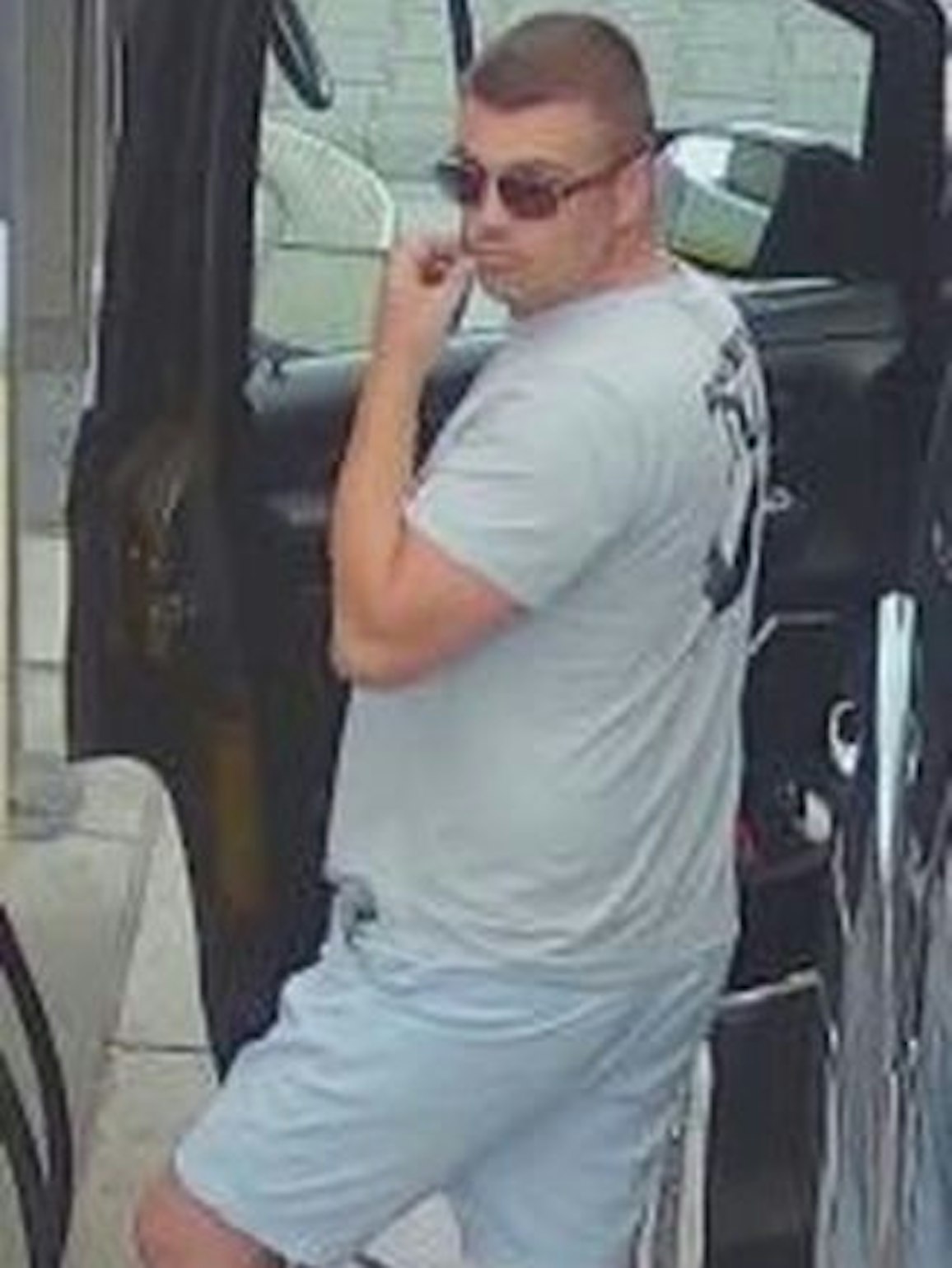 Ein dicklicher Mann in kurzer, hellblauer Hose und grauem T-Shirt steht an einer geöffneten Autotür, er trägt kurz rasierte Haare und eine Sonnenbrille.