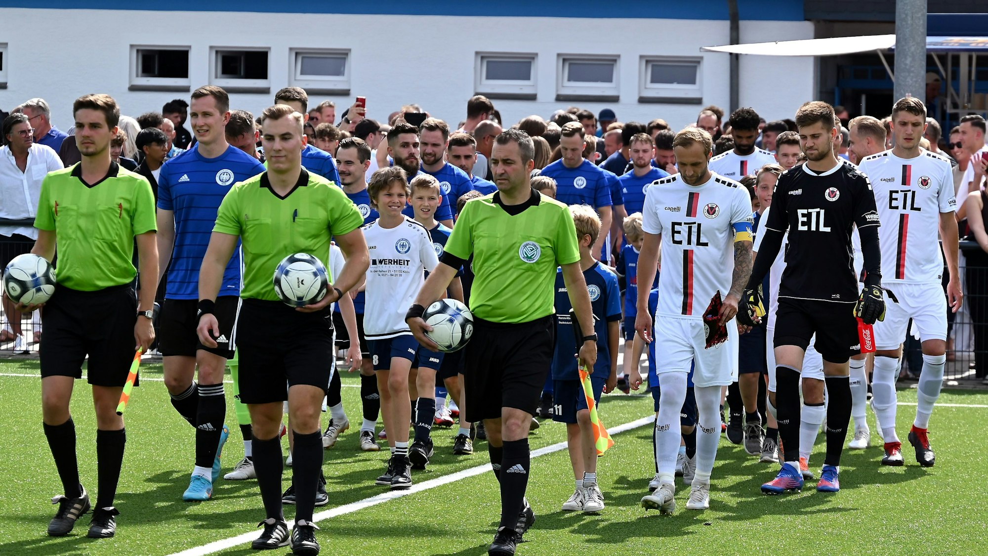 Zwei Fußball-Mannschaften in blauen und weißen Trikots laufen gemeinsam mit den Schiedsrichtern auf das Feld.