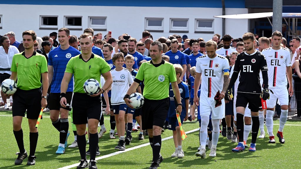 Zwei Fußball-Mannschaften in blauen und weißen Trikots laufen gemeinsam mit den Schiedsrichtern auf das Feld.