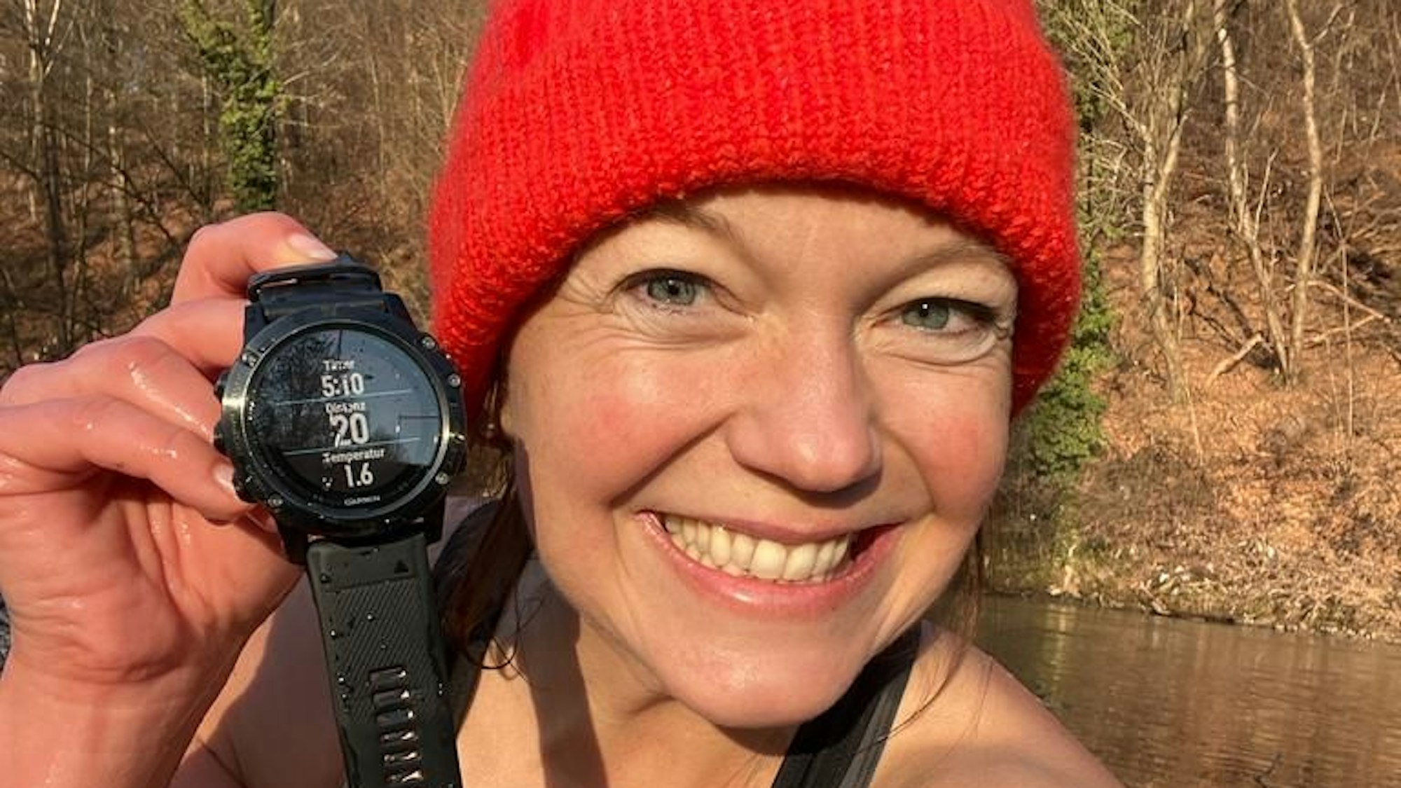 Anita Horn sitzt am Aggerufer in Schwimmkleidung und einer roten Wollmütze. Sie zeigt eine Armbanduhr mit Temperaturanzeige.