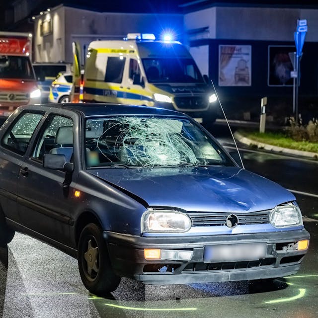 Ein VW-Golf steht mit sichtbaren Unfallspuren auf einer Straße, auf dem Boden sind aufgesprühte Markungen zur Unfallaufnahme zu sehen. Im Hintergrund Einsatzfahrzeuge von Polizei und Rettungsdienst mit eingeschaltetem Blaulicht.
