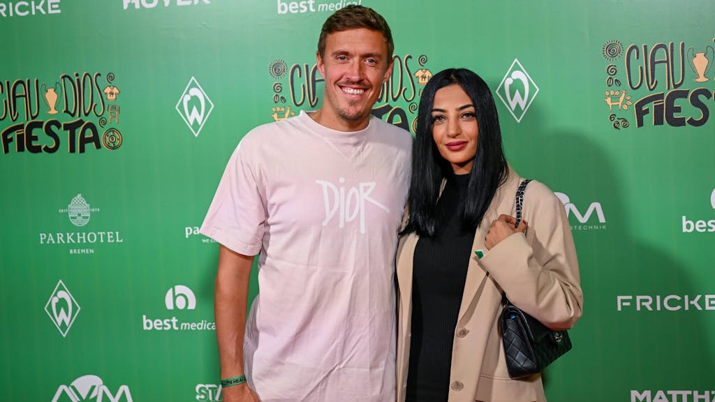 Das Foto zeigt Dilara und Max Kruse im Wohninvest Weserstadion in Bremen vor einer Wand, auf welcher Logos verschiedener Sponsoren des SV Werder Bremen abgedruckt sind.&nbsp;
