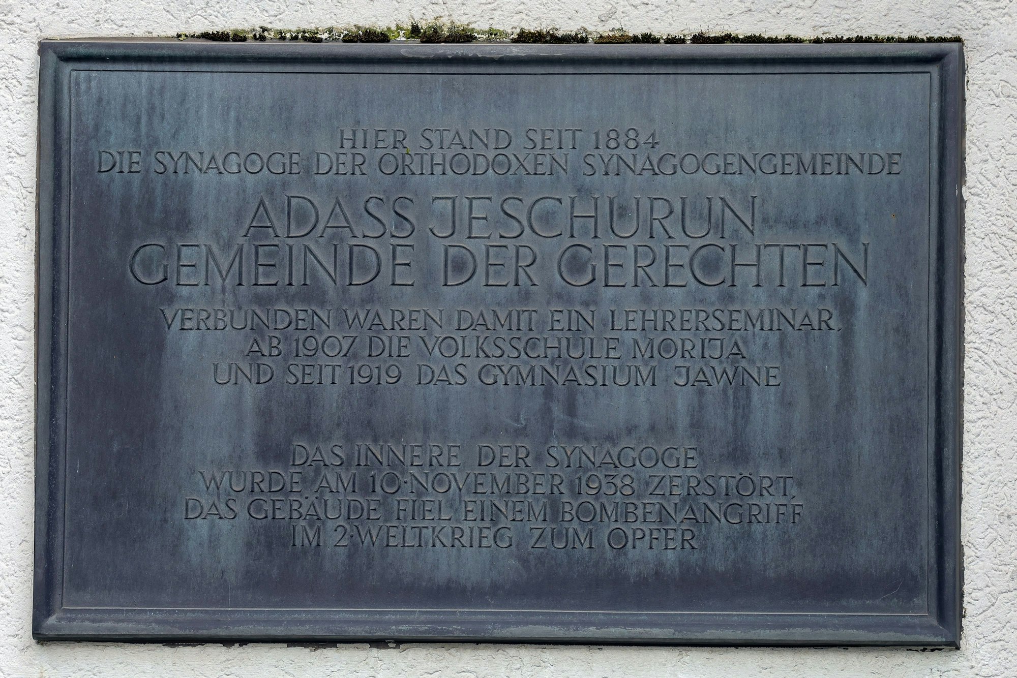 Eine metallene Tafel ist in eine Wand eingelassen. Auf ihr steht neben kleinerer Schrift der Text „Adass Jeschurun \ Gemeinde der Gerechten“.