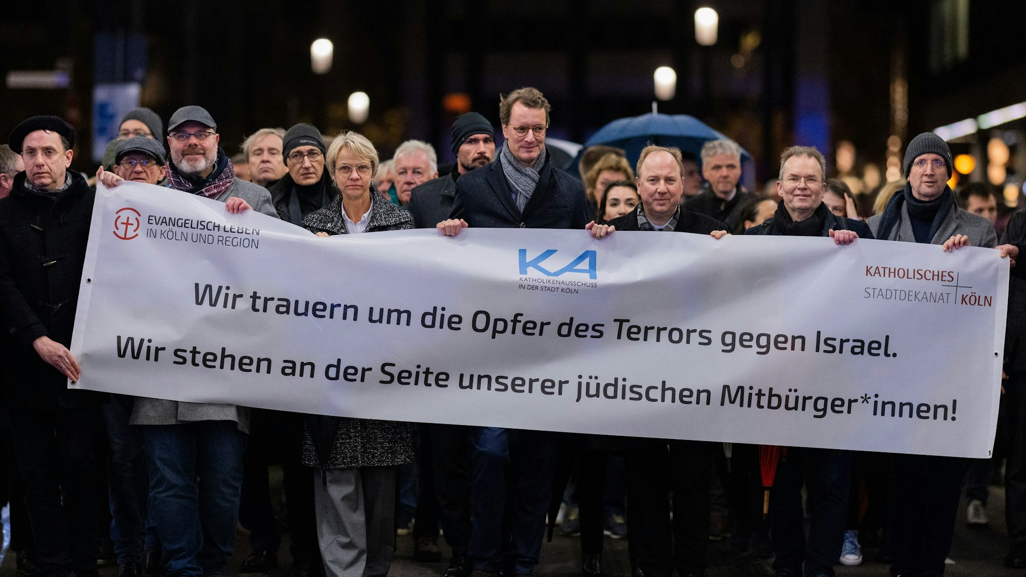 NRW-Ministerpräsident Hendrik Wüst und viele andere Menschen halten ein großes Banner bei einem Schweigegang durch Köln hoch.