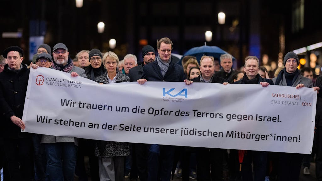 NRW-Ministerpräsident Hendrik Wüst und viele andere Menschen halten ein großes Banner bei einem Schweigegang durch Köln hoch.