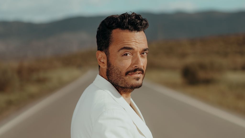 Giovanni Zarrella trägt einen weißen Anzug und blickt auf einer Straße über die Schulter in die Kamera.