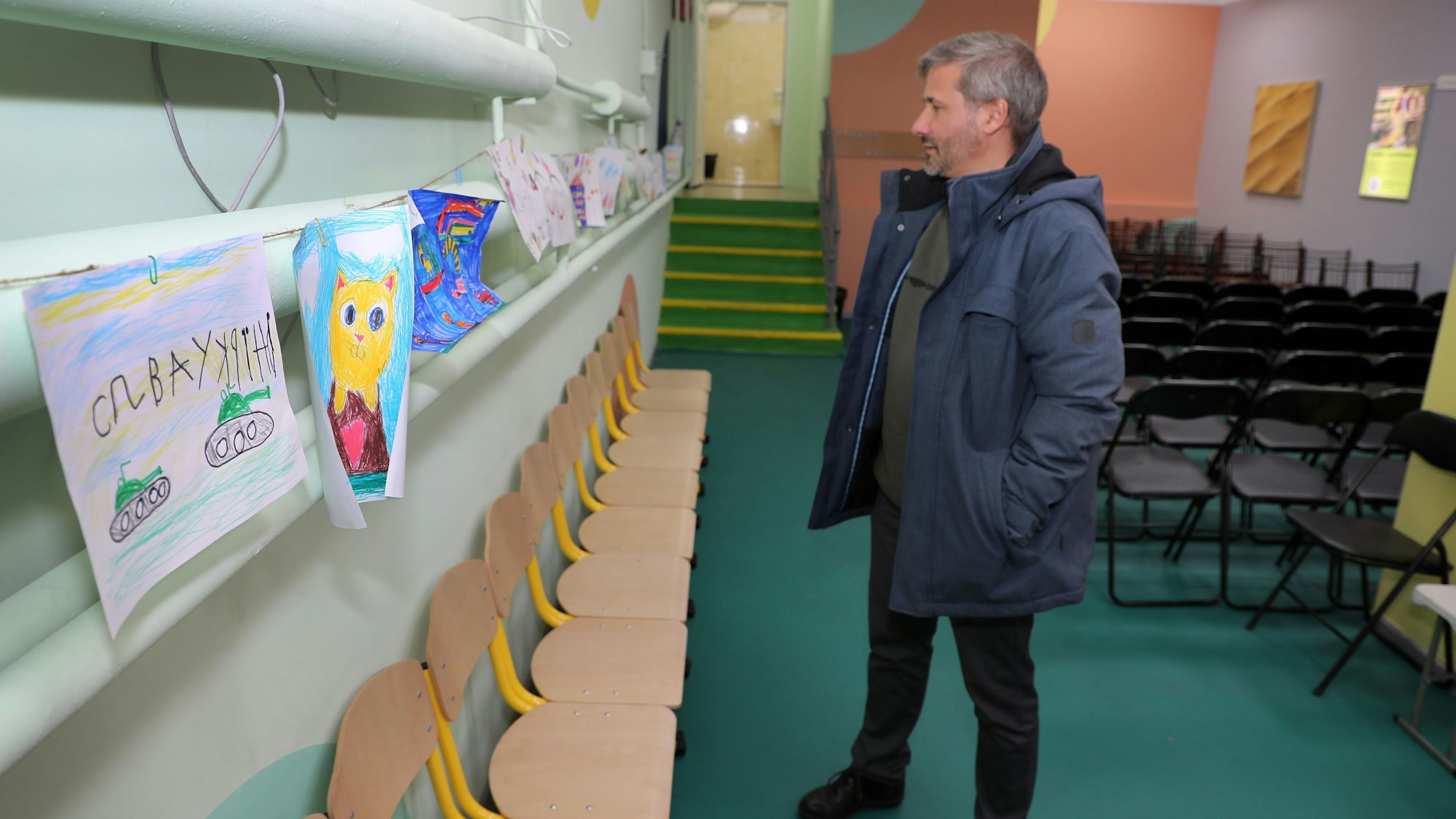 Ein Mann schaut sich Kinderbilder an, die in einem Luftschutzraum unter einer Schule im ukrainischen Butscha an der Wand hängen. Auf einem Bild sind Panzer zu sehen, auf einem anderen eine Katze.