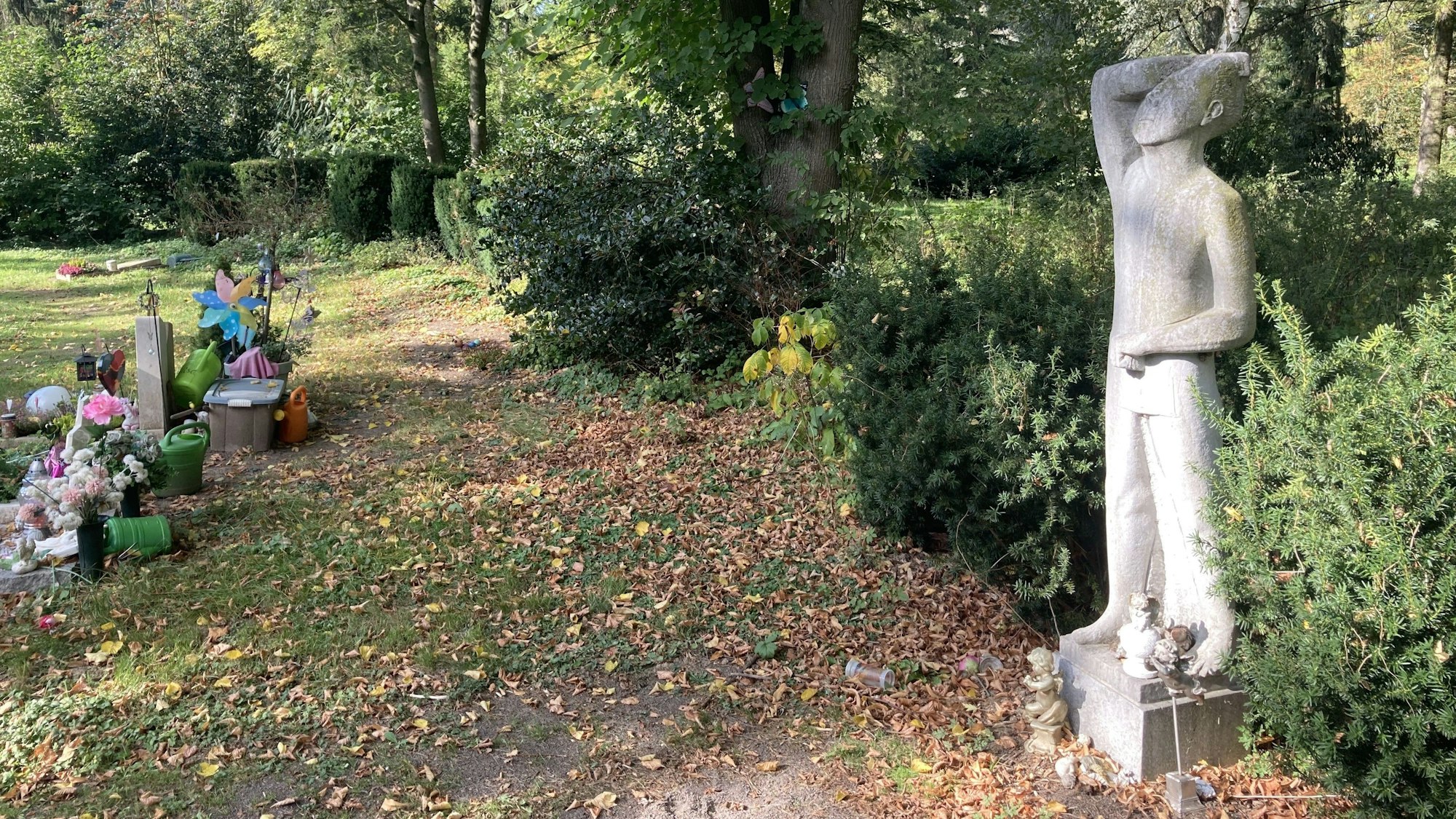 Auf einem Friedhof steht eine einzelne Statue abseits am Gebüsch.