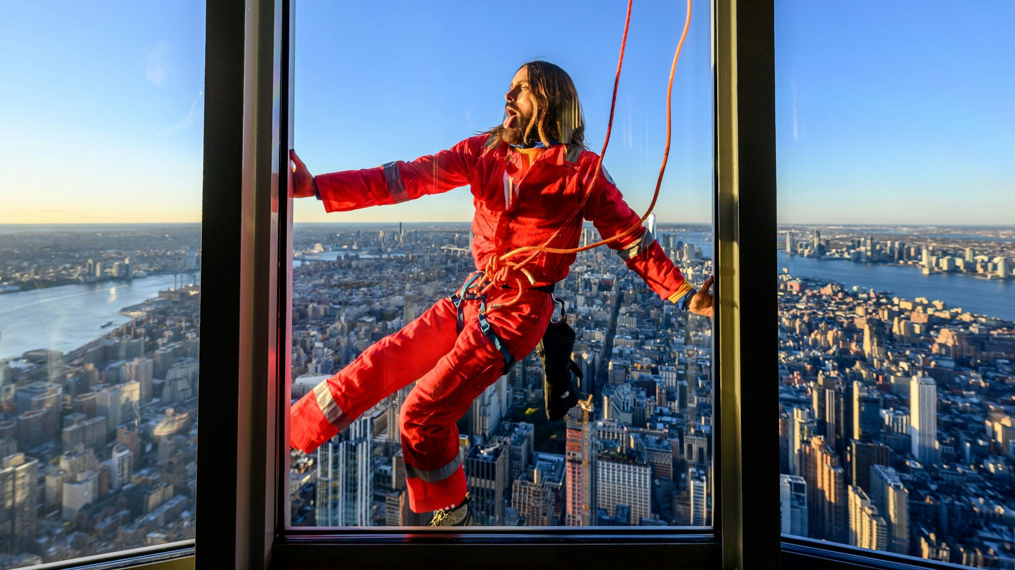 Jared Leto hängt mit einem Seil gesichert am Empire State Building in New York. Er trägt einen roten Ganzkörperanzug.